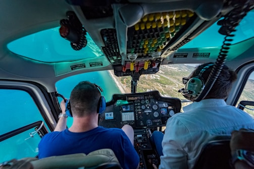 Inside a helicopter cockpit, two pilots are flying over a coastal landscape with clear blue water. The cockpit is filled with instruments and controls, and the pilots are wearing headsets.