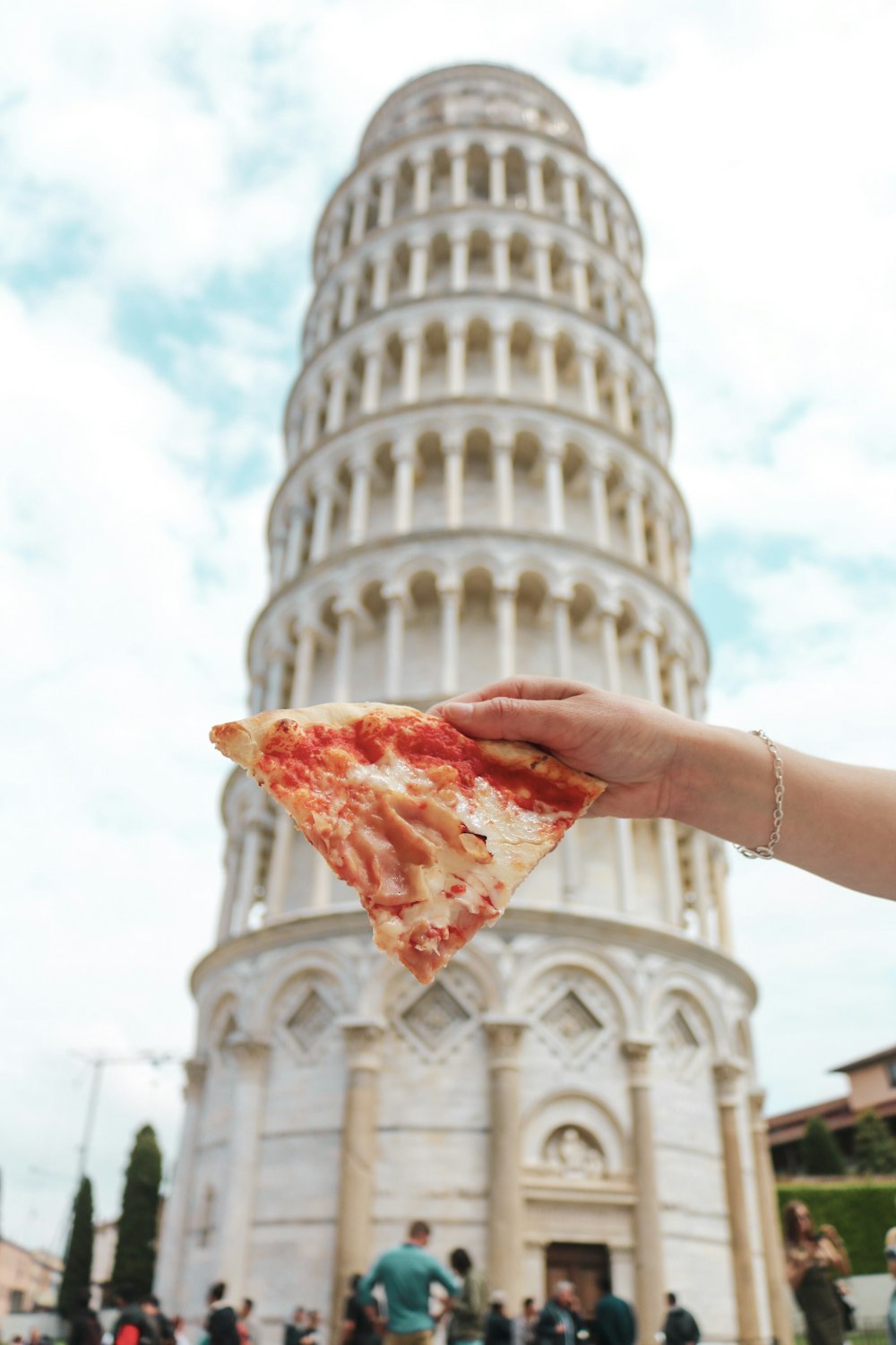 pessoa segurando fatia de pizza em frente à Torre Inclinada de Pisa, Itália durante o dia