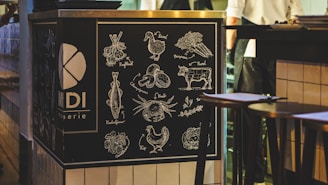 A chalkboard-style illustration on a restaurant counter displays various food items such as vegetables, a cow, chicken, and seafood. Each item is neatly labeled in a decorative manner. The setting is a cozy, dim-lit interior with a hint of activity in the background, likely a restaurant or bistro.