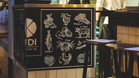 A chalkboard-style illustration on a restaurant counter displays various food items such as vegetables, a cow, chicken, and seafood. Each item is neatly labeled in a decorative manner. The setting is a cozy, dim-lit interior with a hint of activity in the background, likely a restaurant or bistro.