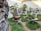 A serene garden display features multiple bonsai trees placed on raised platforms. The bonsais vary in size and shape, each in its own decorative pot, set against a backdrop of white walls and trimmed greenery that covers the ground. The scene is bordered on one side by a large, textured rock formation.