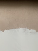 A textured wall with a mix of paint colors, featuring an upper section in beige and a lower section in off-white. Visible brush strokes and uneven painting create a rustic and unfinished look.