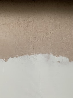 A textured wall with a mix of paint colors, featuring an upper section in beige and a lower section in off-white. Visible brush strokes and uneven painting create a rustic and unfinished look.
