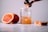 A fresh orange beverage is being poured into a glass jar with a salt rim. The scene includes a half-sliced orange, a whole orange in the background, and a small orange wedge beside the jar on a white surface.