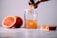 A fresh orange beverage is being poured into a glass jar with a salt rim. The scene includes a half-sliced orange, a whole orange in the background, and a small orange wedge beside the jar on a white surface.