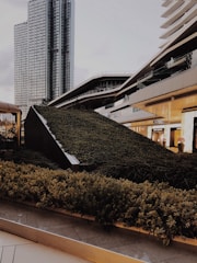 A modern urban landscape featuring a variety of architectural structures, including a tall glass skyscraper and contemporary buildings with sleek, angular designs. The foreground is dominated by a lush green garden that appears to be integrated into the urban design, providing a contrast of nature against the backdrop of city architecture.