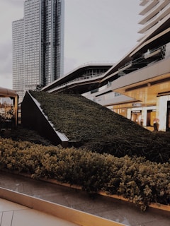 A modern urban landscape featuring a variety of architectural structures, including a tall glass skyscraper and contemporary buildings with sleek, angular designs. The foreground is dominated by a lush green garden that appears to be integrated into the urban design, providing a contrast of nature against the backdrop of city architecture.