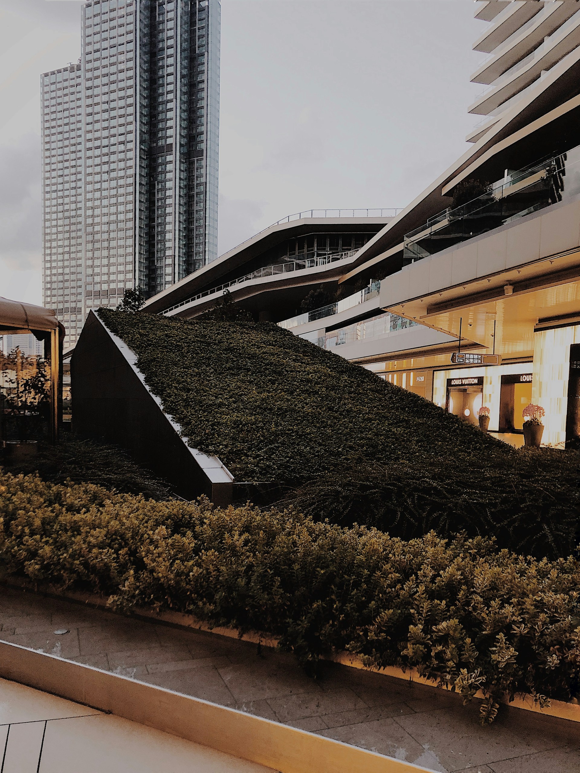A modern urban landscape featuring a variety of architectural structures, including a tall glass skyscraper and contemporary buildings with sleek, angular designs. The foreground is dominated by a lush green garden that appears to be integrated into the urban design, providing a contrast of nature against the backdrop of city architecture.