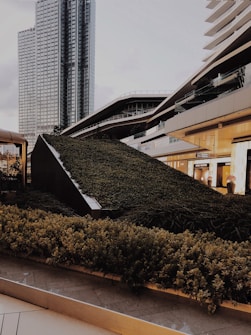 A modern urban landscape featuring a variety of architectural structures, including a tall glass skyscraper and contemporary buildings with sleek, angular designs. The foreground is dominated by a lush green garden that appears to be integrated into the urban design, providing a contrast of nature against the backdrop of city architecture.