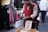 An elderly person wearing a red vest and hat stands in a market setting, handling a plastic bag. A cart with a sign in a foreign script is in front of them. Surrounding them are stalls with hanging clothing and other market goods.