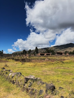 A rural landscape with traditional houses surrounded by open fields and a stone wall in the foreground. The background features rolling hills and tall trees, under a partly cloudy blue sky.