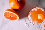 A refreshing orange beverage with ice and a salt rim sits beside whole and sliced oranges on a light-colored surface. The drink garnished with a slice of orange peel creates a vibrant and summery atmosphere.