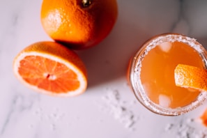 A refreshing orange beverage with ice and a salt rim sits beside whole and sliced oranges on a light-colored surface. The drink garnished with a slice of orange peel creates a vibrant and summery atmosphere.