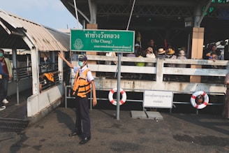 A person in a safety vest and mask stands on a dock next to a green sign with Thai text, including the words 'Tha Chang'. A group of people wearing hats and masks is gathered in the background. Life preservers are attached to the railings, and the dock appears to be part of a ferry terminal.