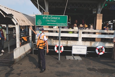 A person in a safety vest and mask stands on a dock next to a green sign with Thai text, including the words 'Tha Chang'. A group of people wearing hats and masks is gathered in the background. Life preservers are attached to the railings, and the dock appears to be part of a ferry terminal.