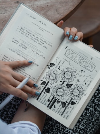 An open journal is held by a person with blue painted nails. The left page contains handwritten notes, including phrases of gratitude and tasks, dated Monday, 19.08.2019. The right page features artistic doodles of flowers labeled with positive traits like 'self love' and 'honesty', surrounded by encouraging words.