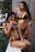 Two women in bikinis are standing and sitting outdoors. They are holding and eating slices of watermelon. One woman is wearing a dark green bikini, and the other is wearing a red bikini. The background includes plant foliage, a tall building structure, and a wooden wall.