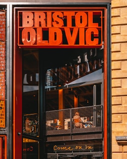 A vibrant red sign displaying 'Bristol Old Vic' is mounted above an entrance. Inside, a person is sitting at a table on a metal balcony with glass and brick elements, signifying a modern and inviting space. A glowing neon sign below reads 'Come on in'.