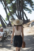 A woman wearing a straw hat and casual summer clothing walks along a shaded path, flanked by tall pine trees. Her back is turned to the camera, and she carries a brown bag. In the background, a man sits at a table, suggesting a relaxed outdoor setting.