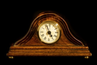 A vintage mantel clock with a polished wooden casing and a round clock face displaying Roman numerals. The background is a stark black, accentuating the warm, rich hues of the wood. The clock hands indicate the time is approximately 10:10.