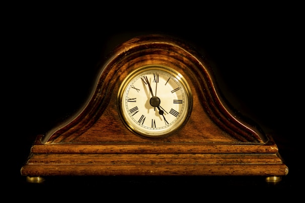A vintage mantel clock with a polished wooden casing and a round clock face displaying Roman numerals. The background is a stark black, accentuating the warm, rich hues of the wood. The clock hands indicate the time is approximately 10:10.