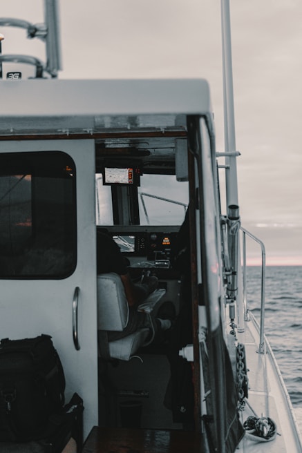 A view of a boat's interior from the doorway, showing the cockpit with an occupant seated at the controls, and marine navigation equipment visible on a panel. The outside ocean and horizon can be seen through the open door.