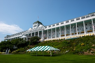 A large, historic hotel with a grand, white facade and numerous windows is situated on a hill. The building features a classic design with tall columns and American flags. The lush greenery and a well-manicured lawn surround the building, with a green and white striped canopy on the lawn.