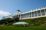 A large, historic hotel with a grand, white facade and numerous windows is situated on a hill. The building features a classic design with tall columns and American flags. The lush greenery and a well-manicured lawn surround the building, with a green and white striped canopy on the lawn.