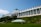 A large, historic hotel with a grand, white facade and numerous windows is situated on a hill. The building features a classic design with tall columns and American flags. The lush greenery and a well-manicured lawn surround the building, with a green and white striped canopy on the lawn.