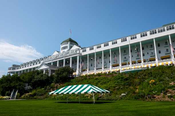 A large, historic hotel with a grand, white facade and numerous windows is situated on a hill. The building features a classic design with tall columns and American flags. The lush greenery and a well-manicured lawn surround the building, with a green and white striped canopy on the lawn.