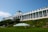 A large, historic hotel with a grand, white facade and numerous windows is situated on a hill. The building features a classic design with tall columns and American flags. The lush greenery and a well-manicured lawn surround the building, with a green and white striped canopy on the lawn.