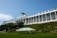 A large, historic hotel with a grand, white facade and numerous windows is situated on a hill. The building features a classic design with tall columns and American flags. The lush greenery and a well-manicured lawn surround the building, with a green and white striped canopy on the lawn.