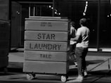 Two individuals wearing casual clothing and caps push large laundry carts in an industrial or urban setting. The carts are labeled 'STAR LAUNDRY TALC' and are mounted on wheels. The background features a glass facade and rows of lights inside the building.
