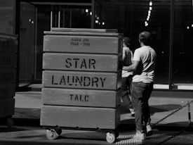 Two individuals wearing casual clothing and caps push large laundry carts in an industrial or urban setting. The carts are labeled 'STAR LAUNDRY TALC' and are mounted on wheels. The background features a glass facade and rows of lights inside the building.