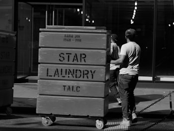 Two individuals wearing casual clothing and caps push large laundry carts in an industrial or urban setting. The carts are labeled 'STAR LAUNDRY TALC' and are mounted on wheels. The background features a glass facade and rows of lights inside the building.