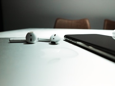 A pair of wireless earphones and a smartphone rest on a reflective white surface, with subtle shadows and a blurred background.