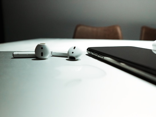 A pair of wireless earphones and a smartphone rest on a reflective white surface, with subtle shadows and a blurred background.