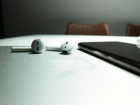 A pair of wireless earphones and a smartphone rest on a reflective white surface, with subtle shadows and a blurred background.