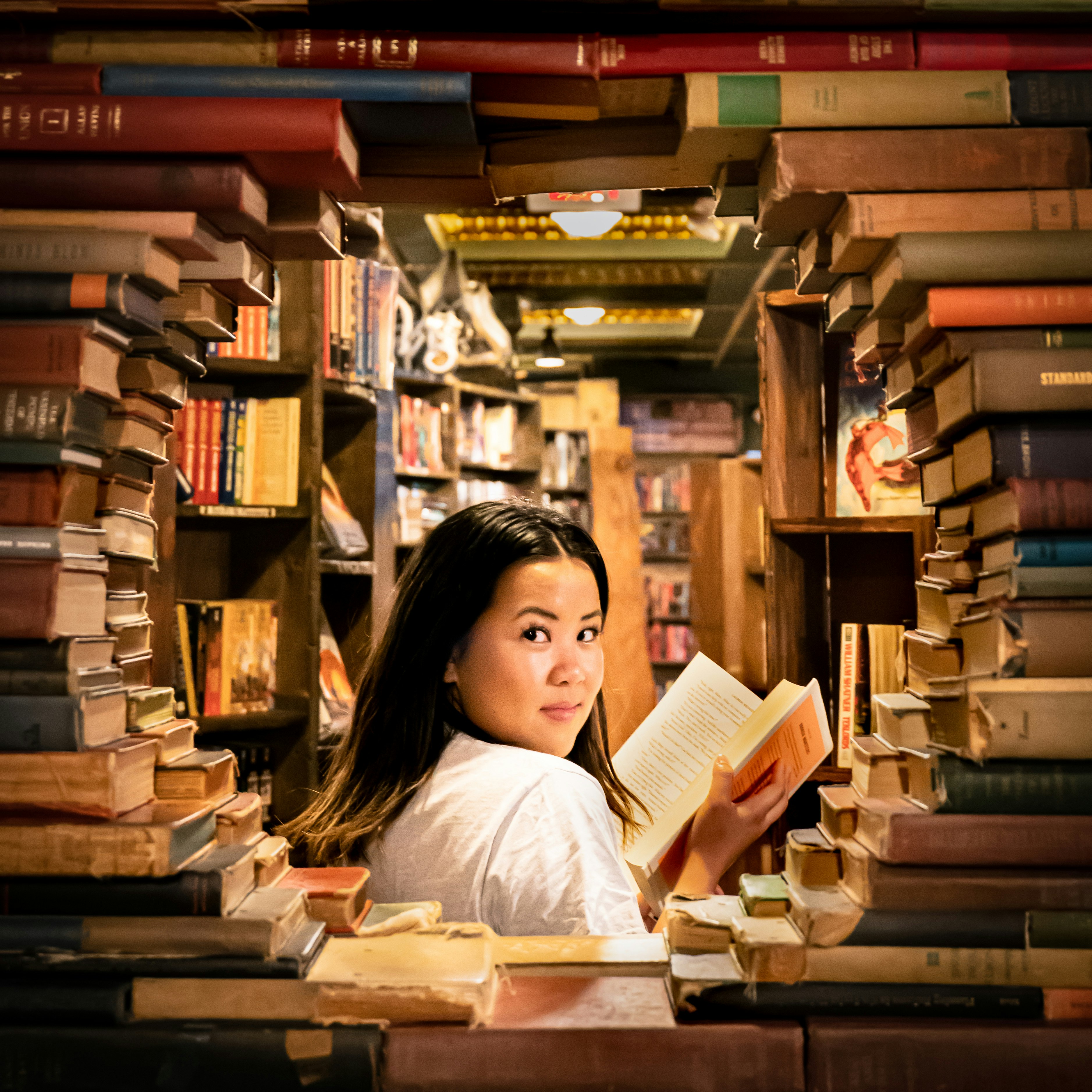 Stock photo of a young woman looking over her shoulder at the viewer, surrounded by books stacked around her