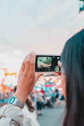 A person is holding a smartphone horizontally, capturing an image of a vibrant, blurred scene likely at a fair or festival. The background is full of colorful lights and people milling around, and the sky is visible above.