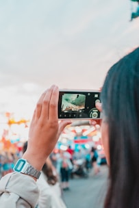 A person is holding a smartphone horizontally, capturing an image of a vibrant, blurred scene likely at a fair or festival. The background is full of colorful lights and people milling around, and the sky is visible above.