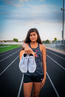 A young woman stands on a running track, holding a pair of white sneakers with orange details directly towards the viewer. She wears athletic clothing, including a sleeveless top with a letter 'L' on it and black shorts. The background features a sports field and a cloudy sky, suggesting a typical outdoor sports environment.