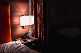 A dimly lit hotel room featuring a nightstand with a digital clock displaying 1:39, a bottle of water, and a smartphone. A modern lamp with a white shade illuminates the area next to an upholstered headboard and a set of drapes with a mix of red and brown hues.
