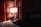 A dimly lit hotel room featuring a nightstand with a digital clock displaying 1:39, a bottle of water, and a smartphone. A modern lamp with a white shade illuminates the area next to an upholstered headboard and a set of drapes with a mix of red and brown hues.