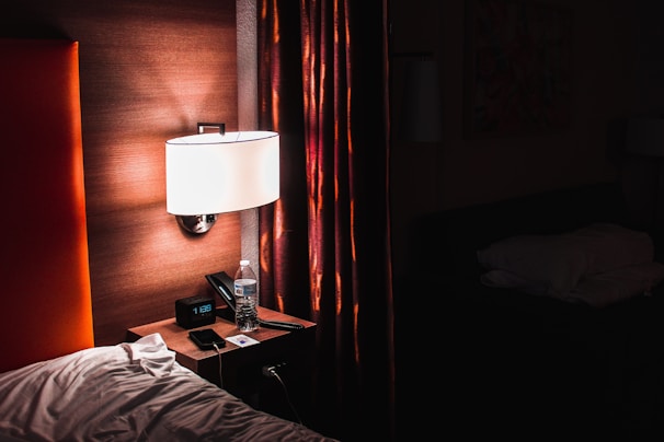 A dimly lit hotel room featuring a nightstand with a digital clock displaying 1:39, a bottle of water, and a smartphone. A modern lamp with a white shade illuminates the area next to an upholstered headboard and a set of drapes with a mix of red and brown hues.