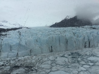 A vast expanse of jagged ice forms a glacier stretching towards distant mountains partially obscured by clouds. The ice surface appears rough and cracked, with various shades of blue and white. Raindrops or condensation are visible on what seems to be a viewing window in the foreground, enhancing the cold and misty ambiance.