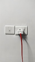 A power outlet on a white wall, featuring two switches and a plugged-in device with a bright red cable extending downward.