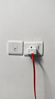 A power outlet on a white wall, featuring two switches and a plugged-in device with a bright red cable extending downward.