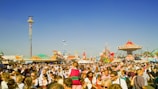 A large crowd of people is gathered at an amusement park or fair, enjoying various rides and attractions. The scene is bustling with activity, featuring a prominent colorful carousel on the right and a tall drop tower ride in the background. Bright and clear blue skies add to the lively atmosphere.