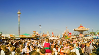 A large crowd of people is gathered at an amusement park or fair, enjoying various rides and attractions. The scene is bustling with activity, featuring a prominent colorful carousel on the right and a tall drop tower ride in the background. Bright and clear blue skies add to the lively atmosphere.